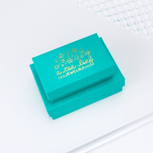 May include: Two turquoise blue boxes stacked on top of each other. The top box has a gold logo that says "a tea leaf jewelry 000".
