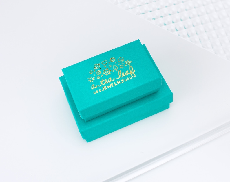 May include: Two turquoise blue boxes stacked on top of each other. The top box has a gold logo that says "a tea leaf jewelry 000".