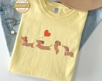 May include: A light yellow Comfort Colors t-shirt featuring a playful design of dachshunds. The dachshunds are brown with various accessories, including a red heart balloon. The shirt is folded on top of blue jeans and a tan hat.