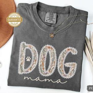 K&ouml;nnte beinhalten: Ein graues Comfort Colors T-Shirt mit den Worten "DOG mama" in einem floralen Design. Das Shirt hat einen Pfotenabdruck im "O" von "DOG". Eine goldene Halskette und ein "Satisfaction Guarantee" Aufkleber sind ebenfalls sichtbar.
