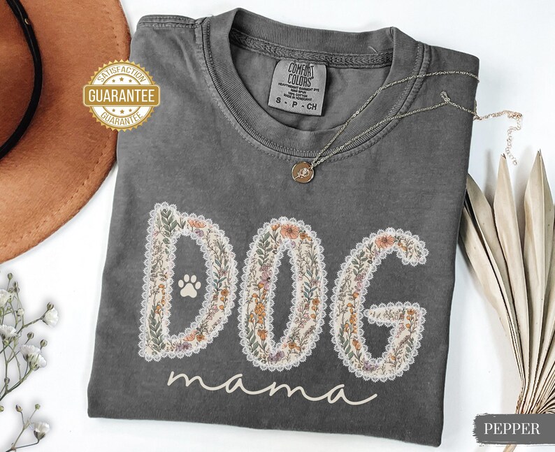 K&ouml;nnte beinhalten: Ein graues Comfort Colors T-Shirt mit den Worten "DOG mama" in einem floralen Design. Das Shirt hat einen Pfotenabdruck im "O" von "DOG". Eine goldene Halskette und ein "Satisfaction Guarantee" Aufkleber sind ebenfalls sichtbar.