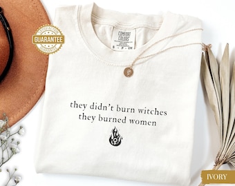Könnte beinhalten: Elfenbeinfarbenes T-Shirt mit dem Text "they didn't burn witches they burned women" in Schwarz. Eine goldene Halskette mit Anhänger liegt auf dem Shirt. Das Shirt hat ein Comfort Colors-Etikett und ein Garantiesiegel.