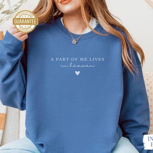 Op de afbeelding: Een blauw sweatshirt met de tekst "A part of me lives in heaven" en een kleine hartvormige afbeelding.