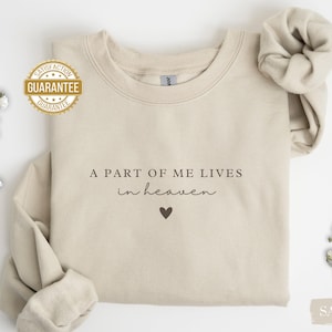 Op de afbeelding: Een beige sweatshirt met de tekst "A part of me lives in heaven" en een klein hartje geprint op de voorkant.