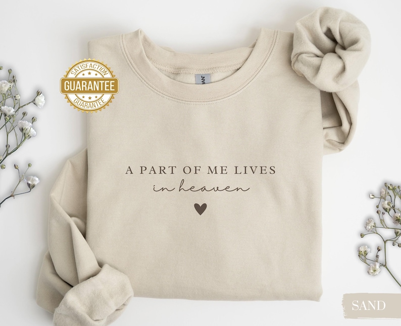 Op de afbeelding: Een beige sweatshirt met de tekst "A part of me lives in heaven" en een klein hartje geprint op de voorkant.