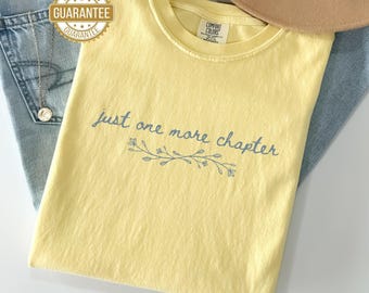May include: A light yellow t-shirt with the text "just one more chapter" in a script font, with a floral design underneath. The shirt is folded on top of a pair of blue jeans. A gold-colored "Satisfaction Guarantee" sticker is visible.