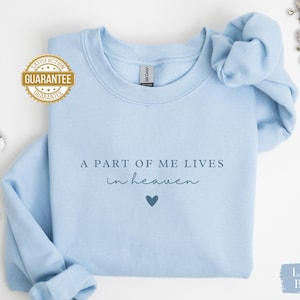Op de afbeelding: Lichtblauwe trui met de tekst "Een deel van mij leeft in de hemel" en een klein blauw hart.