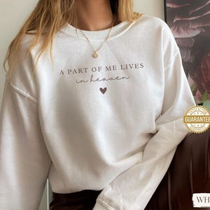 Op de afbeelding: Witte trui met de tekst "A part of me lives in heaven" en een klein hartje.