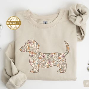 May include: A sand-colored sweatshirt featuring a floral dachshund design. The dog silhouette is filled with a pattern of colorful flowers and foliage. The image also includes a "SATISFACTION GUARANTEE" seal and the word "SAND".