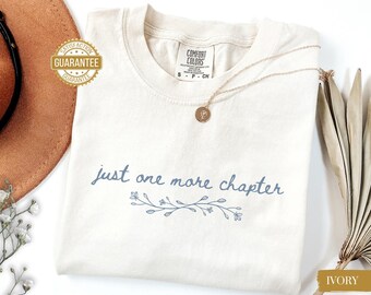 May include: Ivory-colored t-shirt with the text "just one more chapter" in a light blue script, accompanied by a floral design. A gold necklace with a pendant rests on the shirt. The shirt is labeled "Comfort Colors".