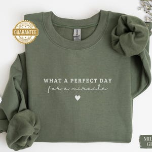 May include: A green sweatshirt with the text "What a perfect day for a miracle" and a small white heart. The sweatshirt is available in military green.