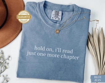 May include: Blue jean colored t-shirt with the text "hold on, I'll read just one more chapter" in white. A gold necklace with a pendant rests on the shirt. The shirt has a crew neck and short sleeves. A "Satisfaction Guarantee" sticker is visible.