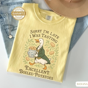 May include: A pale yellow t-shirt with the text "Sorry I'm Late I Was Tasting Excellent Boiled Potatoes." The graphic shows a cartoon duck in a green coat carrying a bowl of potatoes. The shirt is folded on top of blue jeans and a brown hat.