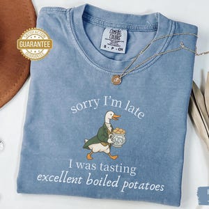 May include: Blue jean-colored t-shirt with the text "sorry I'm late" and "I was tasting excellent boiled potatoes." The graphic features a duck in a green coat carrying a pot of potatoes. A gold necklace rests on the shirt.