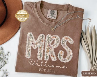 May include: Espresso-coloured t-shirt featuring the word "MRS" in a floral design. The name "Williams" and the year "2025" are printed below. A silver necklace rests on the shirt. The shirt has a Comfort Colors tag.