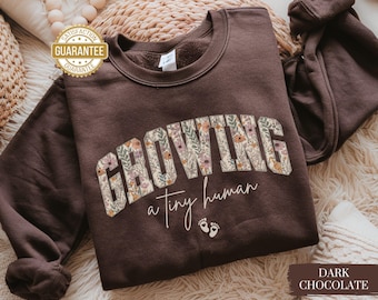 Könnte beinhalten: Dunkelbraunes Sweatshirt mit dem Text "GROWING" in floralem Design und "a tiny human" in Schreibschrift. Mit zwei kleinen Fußabdruck-Grafiken. Ein gemütliches, lässiges Kleidungsstück.