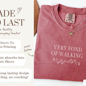 May include: Crimson-colored t-shirt with the text "VERY FOND OF WALKING" in white. The shirt has a crew neck and a small floral design beneath the text. A silver necklace with a pendant rests on the shirt.
