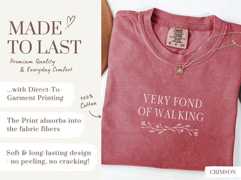 May include: Crimson-colored t-shirt with the text "VERY FOND OF WALKING" in white. The shirt has a crew neck and a small floral design beneath the text. A silver necklace with a pendant rests on the shirt.