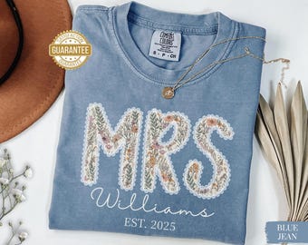 May include: A blue jean coloured t-shirt featuring the word "MRS" in a floral design, the name "Williams", and the year "EST. 2025". A gold necklace with a pendant rests on the shirt. The shirt is labelled "Comfort Colors".
