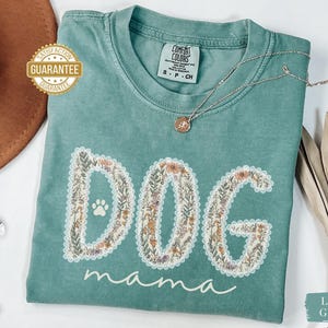 K&ouml;nnte beinhalten: Hellgr&uuml;nes T-Shirt mit den Worten "DOG mama" in einem floralen Design. Das Wort "DOG" zeigt einen Pfotenabdruck. Eine silberne Halskette mit Anh&auml;nger liegt auf dem Shirt. Das Shirt ist mit "Comfort Colors" gekennzeichnet.