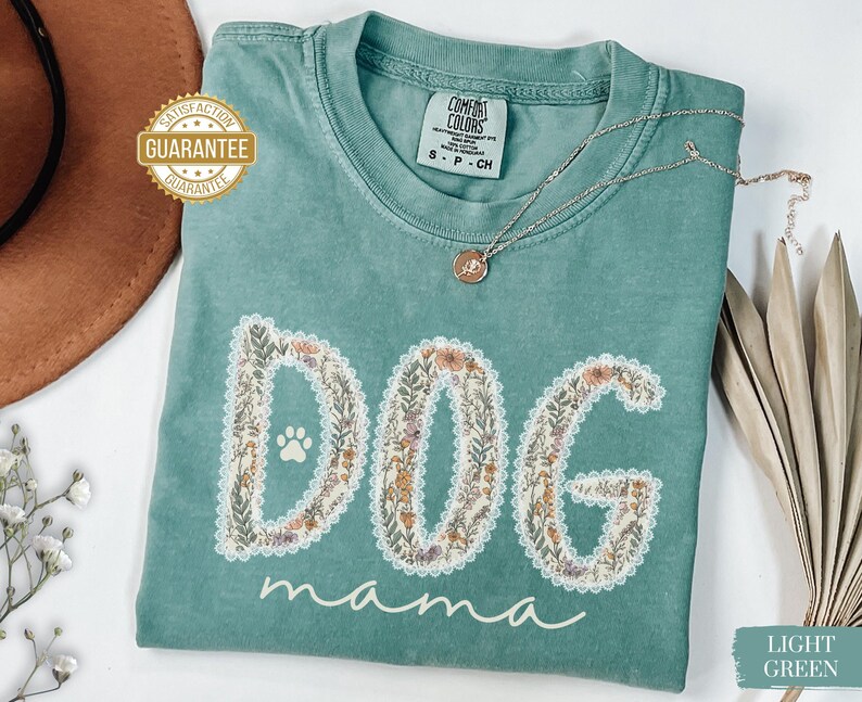 K&ouml;nnte beinhalten: Hellgr&uuml;nes T-Shirt mit den Worten "DOG mama" in einem floralen Design. Das Wort "DOG" zeigt einen Pfotenabdruck. Eine silberne Halskette mit Anh&auml;nger liegt auf dem Shirt. Das Shirt ist mit "Comfort Colors" gekennzeichnet.