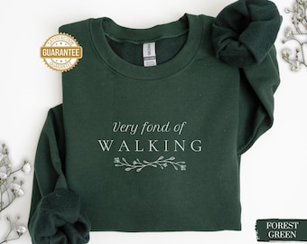Könnte beinhalten: Forstgrünes Sweatshirt mit dem Text "Very fond of WALKING" in weißer, eleganter Schrift. Unter dem Text befindet sich ein florales Design. Das Sweatshirt hat einen Rundhalsausschnitt und lange Ärmel. Ein goldener Garantiesiegel befindet sich oben links.