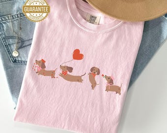 May include: A light pink t-shirt featuring a line of dachshunds with accessories, including a heart-shaped balloon and a rose. The shirt is labeled "BLOSSOM" at the bottom and has a "Satisfaction Guarantee" seal.