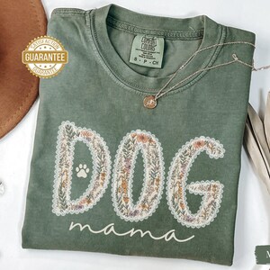 K&ouml;nnte beinhalten: Moosgr&uuml;nes T-Shirt mit den Worten "DOG mama" in einem floralen Design. Das Wort "DOG" zeigt einen Pfotenabdruck. Eine goldene Halskette mit Anh&auml;nger liegt auf dem Shirt. Das Shirt ist aus weichem Material.