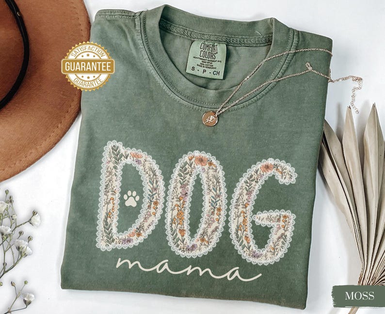 K&ouml;nnte beinhalten: Moosgr&uuml;nes T-Shirt mit den Worten "DOG mama" in einem floralen Design. Das Wort "DOG" zeigt einen Pfotenabdruck. Eine goldene Halskette mit Anh&auml;nger liegt auf dem Shirt. Das Shirt ist aus weichem Material.
