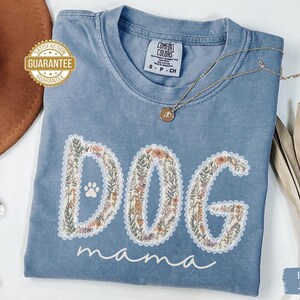 K&ouml;nnte beinhalten: Ein blaues Comfort Colors T-Shirt mit dem Schriftzug "DOG mama" in einem floralen Design. Das Shirt hat einen Rundhalsausschnitt und kurze &Auml;rmel. Eine goldene Halskette mit Anh&auml;nger liegt &uuml;ber dem Shirt.