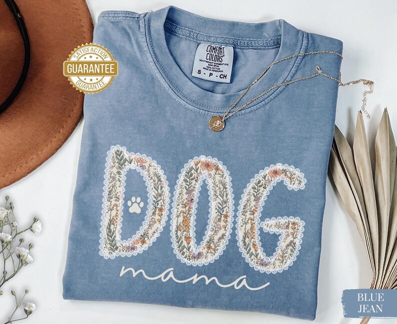 K&ouml;nnte beinhalten: Ein blaues Comfort Colors T-Shirt mit dem Schriftzug "DOG mama" in einem floralen Design. Das Shirt hat einen Rundhalsausschnitt und kurze &Auml;rmel. Eine goldene Halskette mit Anh&auml;nger liegt &uuml;ber dem Shirt.