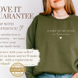 Op de afbeelding: Een groen sweatshirt met de tekst "A part of me lives in heaven" en een klein hartje. De tekst is wit en het hartje is roze. Het sweatshirt wordt gedragen door iemand die een blauwe jeans draagt.