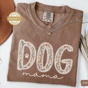 K&ouml;nnte beinhalten: Espressofarbenes T-Shirt mit den Worten "DOG mama" in einem floralen Design. Das Wort "DOG" hat einen Pfotenabdruck. Eine silberne Halskette mit Anh&auml;nger liegt auf dem Shirt. Das Shirt hat ein Comfort Colors-Etikett.