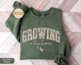 Könnte beinhalten: Ein militärgrünes Sweatshirt mit dem Wort "GROWING" in floralem Design auf der Brust. Darunter steht der Text "a tiny human" mit zwei kleinen Fußabdrücken. Das Sweatshirt hat einen Rundhalsausschnitt und lange Ärmel.