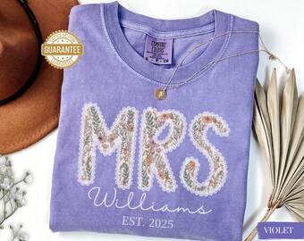 May include: A violet Comfort Colors t-shirt featuring the word "MRS" in a floral design, the name "Williams", and the year "EST. 2025". A gold necklace and a brown hat are also visible.