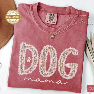 K&ouml;nnte beinhalten: Karmesinrotes T-Shirt mit den Worten "DOG mama" in einem floralen Design. Das Wort "DOG" hat einen Pfotenabdruck. Eine silberne Halskette mit Anh&auml;nger liegt auf dem Shirt. Das Shirt hat ein Comfort Colors-Etikett.
