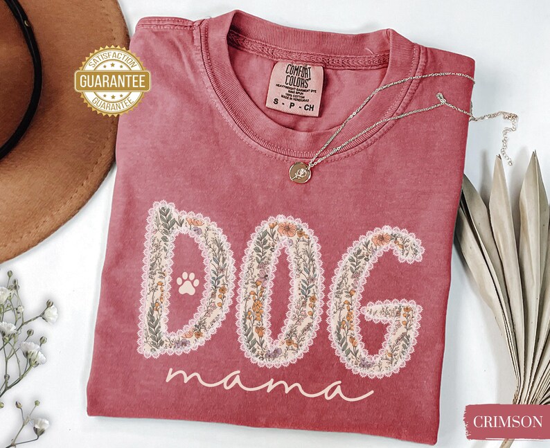 K&ouml;nnte beinhalten: Karmesinrotes T-Shirt mit den Worten "DOG mama" in einem floralen Design. Das Wort "DOG" hat einen Pfotenabdruck. Eine silberne Halskette mit Anh&auml;nger liegt auf dem Shirt. Das Shirt hat ein Comfort Colors-Etikett.