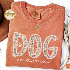 K&ouml;nnte beinhalten: Ein korallenfarbenes T-Shirt mit den Worten "DOG mama" in einem floralen Design. Das Wort "DOG" hat einen Pfotenabdruck in der Mitte. Eine goldene Halskette und ein brauner Hut sind im Hintergrund zu sehen.