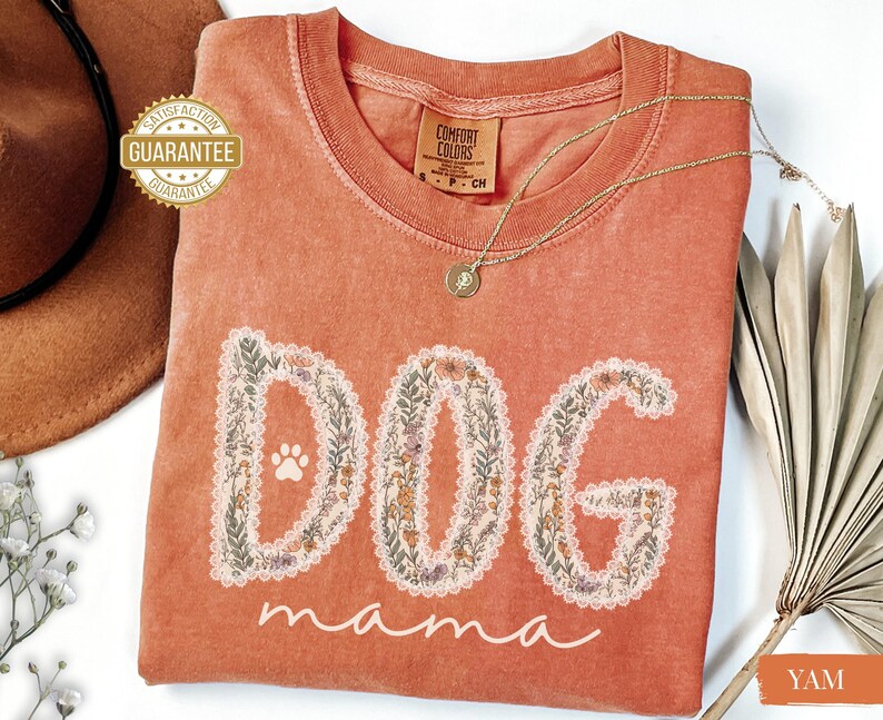 K&ouml;nnte beinhalten: Ein korallenfarbenes T-Shirt mit den Worten "DOG mama" in einem floralen Design. Das Wort "DOG" hat einen Pfotenabdruck in der Mitte. Eine goldene Halskette und ein brauner Hut sind im Hintergrund zu sehen.