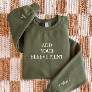 May include: Olive green sweatshirt with the text "ADD YOUR SLEEVE PRINT" in white. The sleeves have the word "Name" printed on them. The sweatshirt is displayed on a brown and white patterned surface.