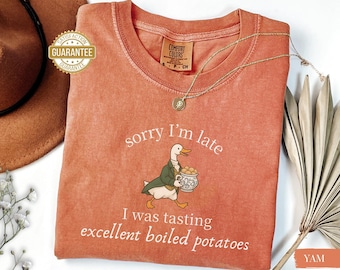 Puede incluir: Una camiseta de color terracota con el texto "sorry I'm late" y "I was tasting excellent boiled potatoes". Un pato de dibujos animados con un abrigo verde lleva una olla de patatas. También se ven un collar dorado y un sombrero marrón.
