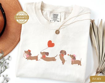 May include: Ivory-colored t-shirt featuring a Comfort Colors label and a graphic of four dachshunds. The dachshunds are wearing accessories and holding a heart-shaped balloon and a rose. A gold necklace is also visible.