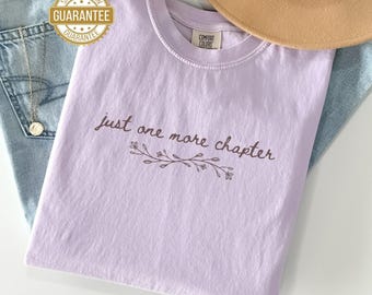 May include: A light purple t-shirt with the text "just one more chapter" in a brown script font, with a floral design underneath. The shirt is folded and rests on a pair of blue jeans. A tan hat is visible in the background.