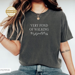 May include: Dark gray t-shirt with the text "VERY FOND OF WALKING" in white, along with a floral design. The shirt has a vintage, worn look. The word "PEPPER" is at the bottom of the image.