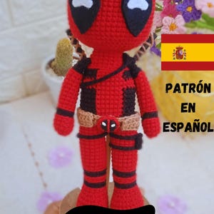 Könnte beinhalten: Eine rot-schwarze gehäkelte Deadpool Amigurumi-Puppe mit einem braunen Gürtel. Die Puppe steht auf einem Holzsockel mit dem Wort "DEADPOOL" darauf. Der Hintergrund zeigt einen Kaktus und Blumen.