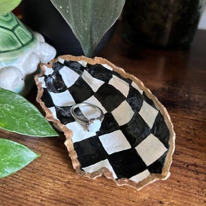 May include: A black and white chequered ring dish made from a seashell with a gold rim. The dish is sitting on a wooden surface and a ring is resting in the centre.