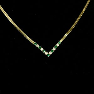 May include: A gold-toned necklace with a V-shaped pendant. The pendant is adorned with alternating green and clear gemstones. The necklace has a herringbone chain and is set against a black background.
