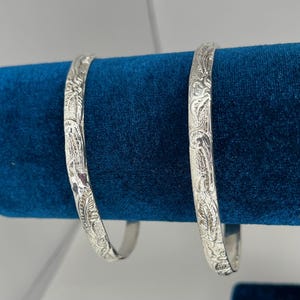 May include: Two silver bangles with intricate floral patterns are displayed against a deep blue velvet backdrop. The bangles are round and have a detailed, engraved design. The light reflects off the polished metal, highlighting the delicate carvings.