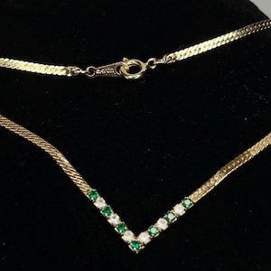May include: Gold-toned necklace with a V-shaped pendant. The pendant is adorned with alternating green and clear gemstones. The necklace has a herringbone chain and a clasp marked "AVON".