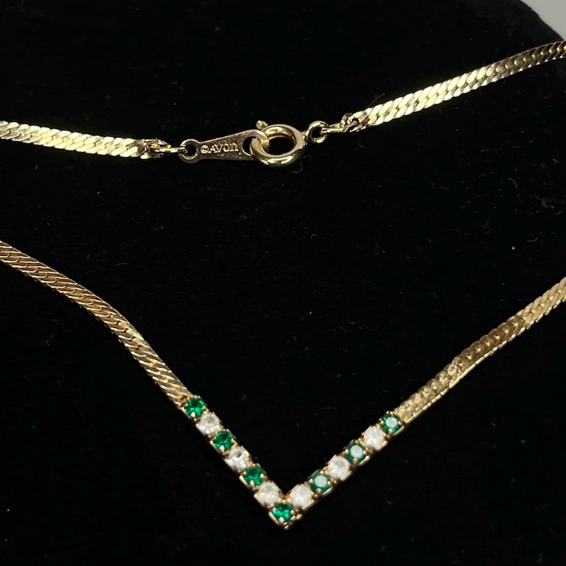 May include: Gold-toned necklace with a V-shaped pendant. The pendant is adorned with alternating green and clear gemstones. The necklace has a herringbone chain and a clasp marked "AVON".